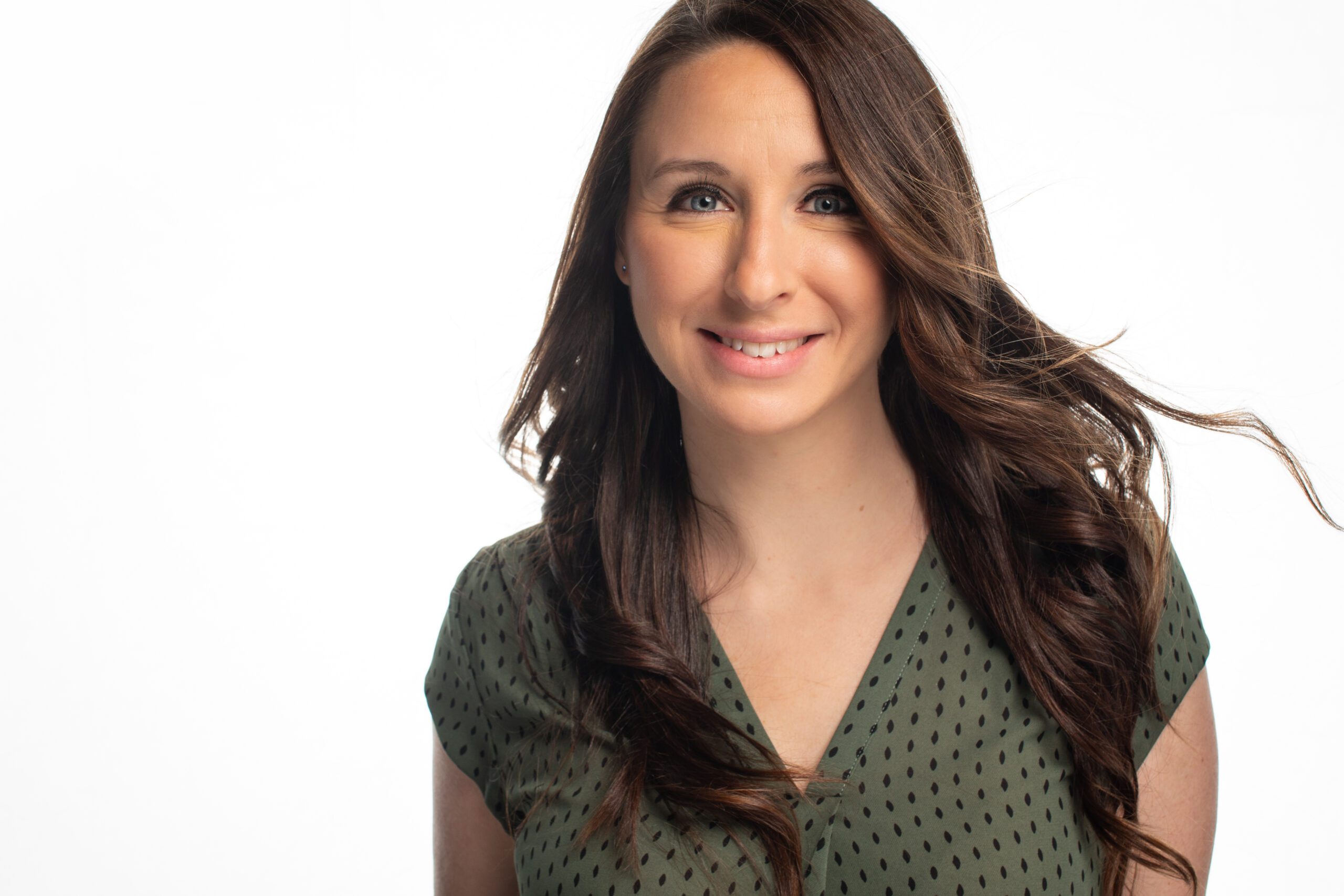 IMG_0305 Woman with long brown hair smiling, wearing a green patterned top, against a white background.